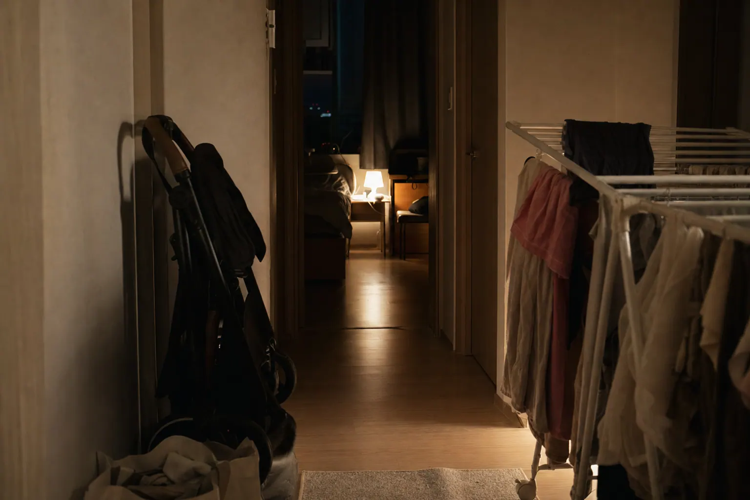 A narrow apartment hallway at night, a stroller folded against the wall, a single lamp left on in a bedroom doorway, laundry drying on a rack, everything quiet and slightly out of order.