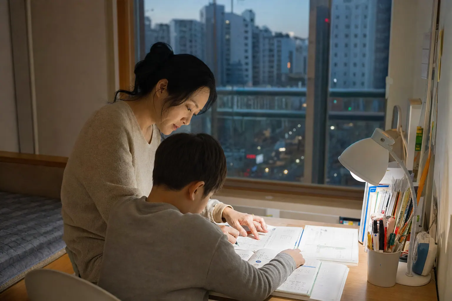 A photorealistic documentary photograph of a Korean mother helping a school-age child with homework at a small apartment desk in evening, textbooks open, a window showing a cold city dusk outside.