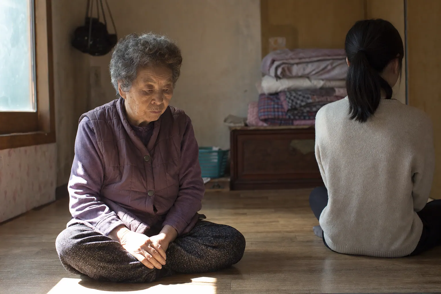 A photorealistic documentary photograph of an elderly Korean woman sitting on the wooden floor of a small village home, sunlight through a window falling on her hands folded in her lap, a younger person sitting slightly apart with their back partly turned, warm dim interior with folded blankets in the corner.
