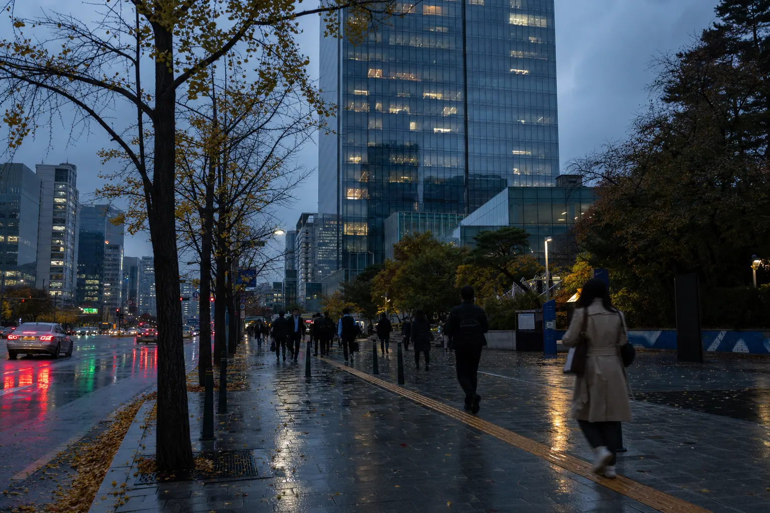 A photorealistic documentary photograph of a glass office tower in Seoul at dusk, most windows dark and a few still lit, seen from street level with commuters walking home along a wet pavement, umbrellas closed, autumn leaves gathered near the curb.