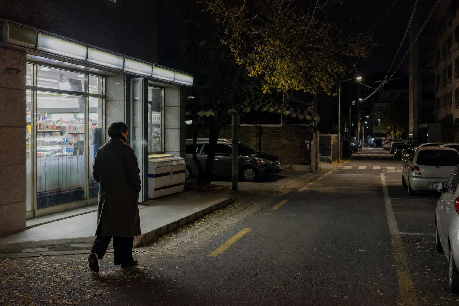 A photorealistic documentary photograph of a person in a long coat walking alone past a lit convenience store on a quiet Seoul side street after dark in late autumn, faint breath visible in the cold air.